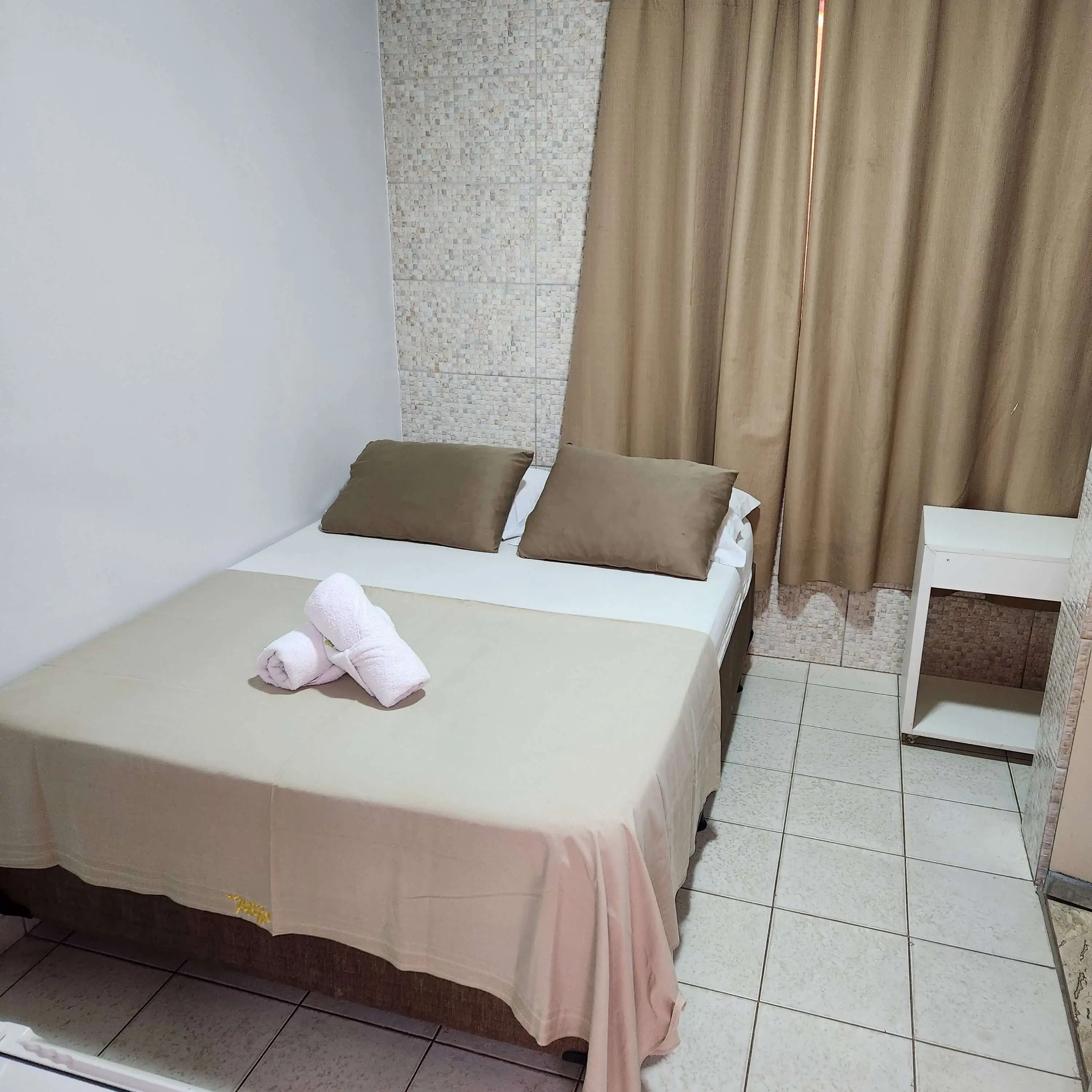 quarto 125 do hotel, com piso branco, cortinas douradas, cama de casal, masa para colocar objetos pessoais, e 2 toalhas com sabonetinho em cima da cama.